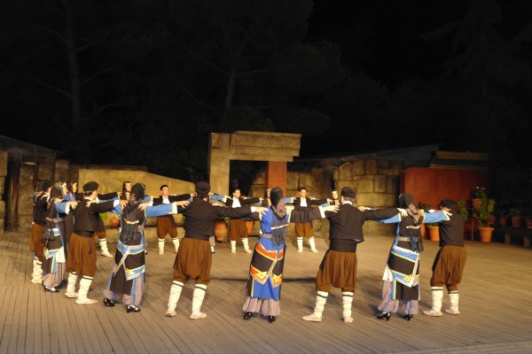 Women and men in traditional attire are dancing in a circle on a big stage.