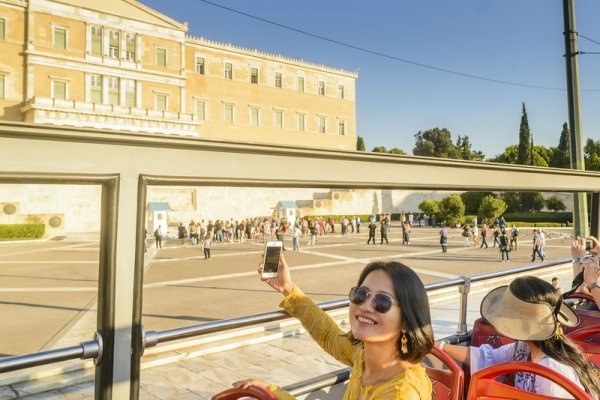 A woman is posing with her phone in hand in front of the Greek parliament on the top deck of a hop on hop off bus.