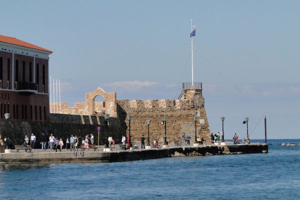 The brown-stone walls of the Firka Venetian Fortress by the sea and tourists taking a walk by. 