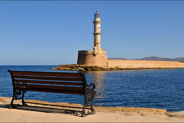 A bench by the deep-blue sea with a view of the Chania's Lighthouse, which is built with light-brown stones.