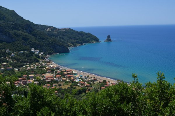 Agios Gordios beach and the rock in the sea at the feet of a massive hill with forest.