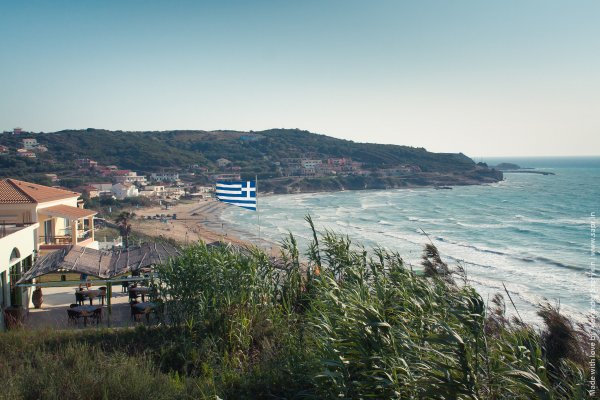 A tavern, a house and the beach and sea of Arillas under a Greek flag.