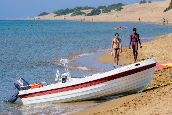 Two women walk in front of a boat with sand dunes in the background at Issos Beach, Ag. Georgios.