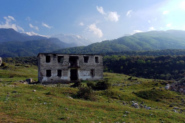 An abandoned building at Old Vrontou Village in a green landscape with high mountains in the background.