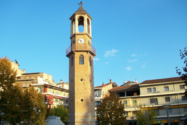 The clock tower of Grevena having in the background blocks of apartments and a blue sky.