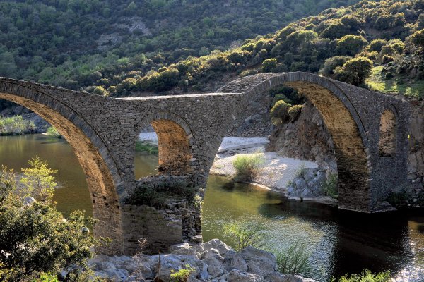 The arched stone bridge over Kompsatos river and hills covered by bushes in the background.