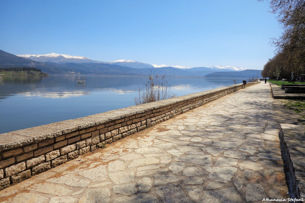 A picture showing the lakeshore promenade, the lake itself and the surrounding mountains of Ioannina Lake.