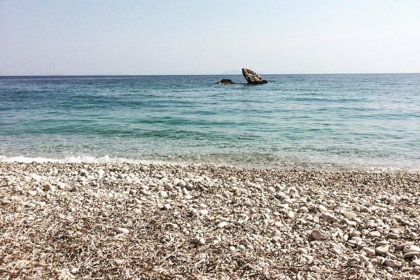 Pebbles, turquoise waters, a rock in the sea, and the blue sky of Ragia, Poros.