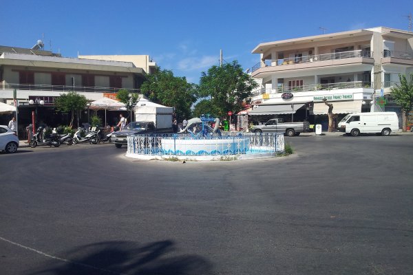 A white-blue circular fountain with dolphins and apartments with parked cars and motorbikes in front of them.