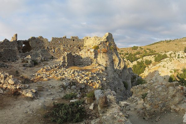 Ruined walls and fallen stones in an arid landscape with bushes and sparse vegetation.