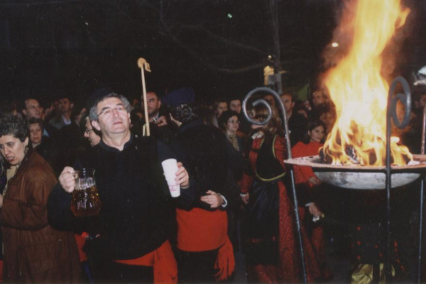 People in traditional clothes, holding a jug of wine, are singing around a lit metal cresset.