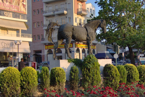 The statue of Bucephalus surrounded by bushes and flowers, having some blocks of flats of Larissa in the background.