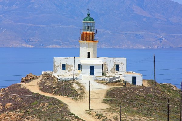 A white lighthouse with a green dome on top of a hill with land and the sea in the background. 