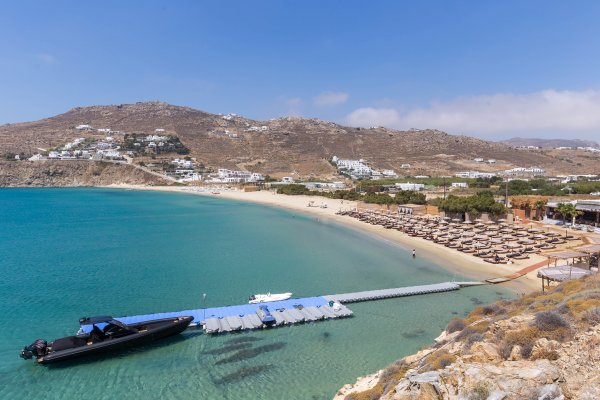 An aerial view of Kalo Livadi Beach, Mykonos, with a small floating pier, a boat, and a long beach with only one beach bar. 