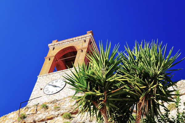 The Clock of Nafpaktos built of brick and stone, and a palm tree against a blue sky.