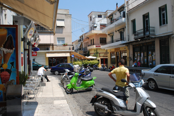 A street with two-story residencies a cafeteria and parked motorbikes just before the Stenopazaro alley.