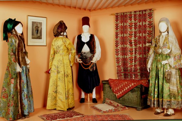 Four mannequins in traditional green, yellow, and black attire in an orange room in the Foundation V. Papantoniou.