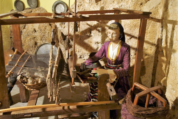 A representation of a woman working at an old wooden loom in a picture where earth colors dominate.