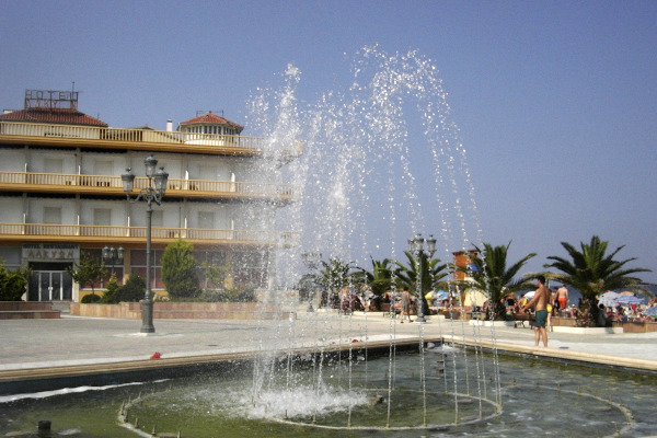 The fountain of Paralia at the square by the beach and short palm trees.