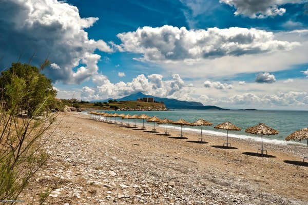 A line of beach umbrellas by the sea and bushes, under a cloudy sky at Pythagorion, Samos.