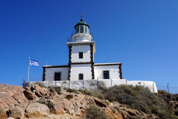 A white lighthouse with brown-rock corners that shape squares sits atop a rocky hill and a Greek flag.