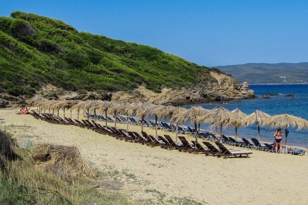 Lined-up beach umbrellas and sunbeds with a green and rocky hill in the background, at Mandraki Beach, Skiathos.