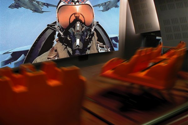 A fighter jet pilot on the screen of the simulator room and seats thrusting forward.
