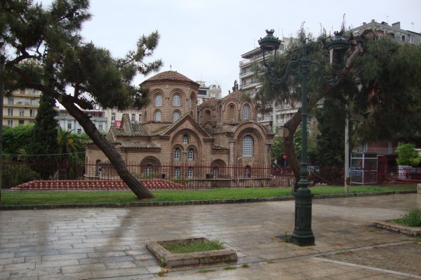 The brick-built Byzantine Church of Panagia Chalkeon behind the paved park and a few trees, surrounded by block of apartments.