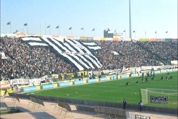 Thousands of PAOK fans hold up black and white banners and a huge football t-shirt.
